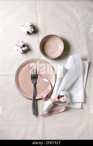 Set of empty pink ceramic plates with fork and textile napkin, decorated by cotton flowers and silk ribbon over white linen tablecloth. Table setting. Stock Photo