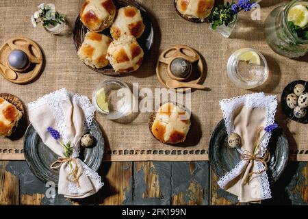 Easter table setting with colored and chocolate eggs, hot cross buns, bouquet flowers, empty ceramic plate with napkin, glass of lemonade drink on woo Stock Photo