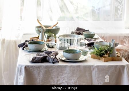 Rustic Christmas table setting with empty craft ceramic tableware, plates and bowls, xmas angel decorations, glasses, green branches on white tableclo Stock Photo