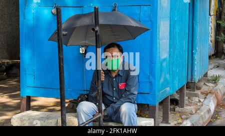 Kolkata, India. 27th Apr, 2021. A man wearing a mask, sitting on a rock holding umbrella and waiting for the vehicle. West Bengal Government has confirmed 16000 COVID-19 cases so far with 9775 recoveries and 68  deaths at Tuesday, 27/04/2021. (Photo by Anirban Lahiri/Pacific Press) Credit: Pacific Press Media Production Corp./Alamy Live News Stock Photo