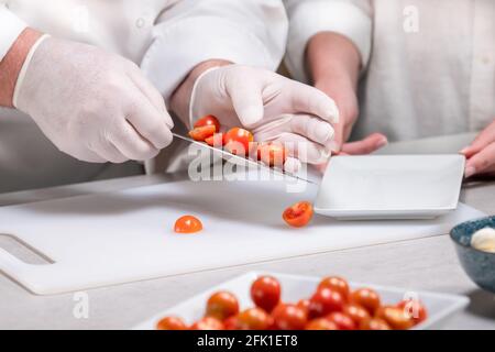 Male chef wearing gloves putting cut cherry tomatoes on a dish using his knife: selective focus Stock Photo