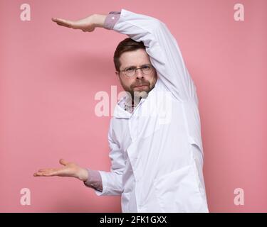 Serious doctor in round glasses shows something in a strange pose and looks sternly at the camera. Copy space. Pink background. Stock Photo