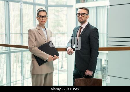 Portrait of serious colleagues in formal wear standing at glassy railing and leaning on it in business hall Stock Photo