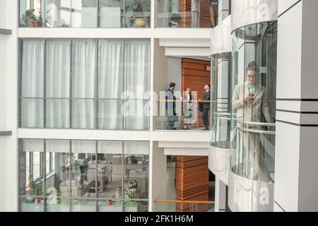 Busy people working in business hall: three colleagues discussing project, young woman checking phone in glassy elevator Stock Photo