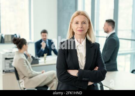 Portrait of content mature blond-haired lady in black jacket standing with crossed arms in business office with colleagues in background Stock Photo