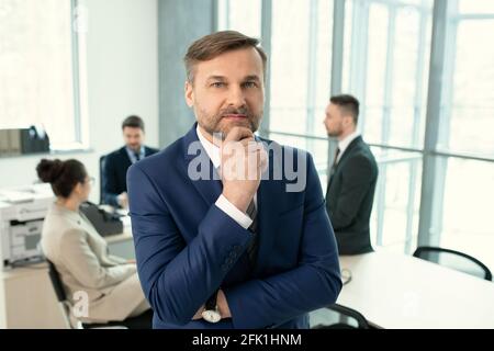 Portrait of pensive handsome mature executive in black suit touching chin while standing in meeting room full of business colleagues Stock Photo