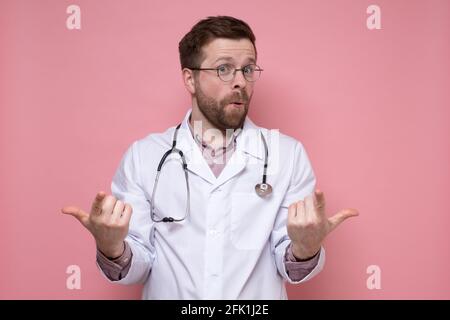 Satisfied, cute doctor with a stethoscope around neck makes a gesture with hands and fingers and looks at the camera in surprise.  Stock Photo