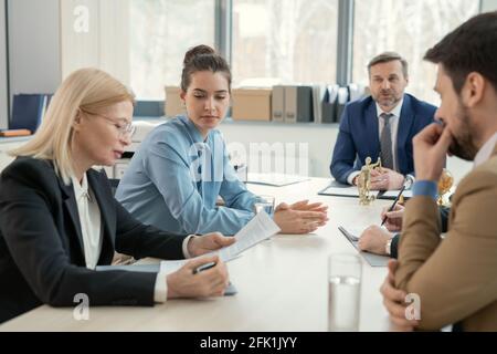 Concentrated mature lawyer sitting at table and analyzing document at meeting with opponents of her client in presence of judge Stock Photo