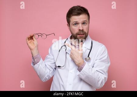 Doctor holds glasses in hands and points at them with finger, explaining that he cannot live without lenses due to poor eyesight.  Stock Photo