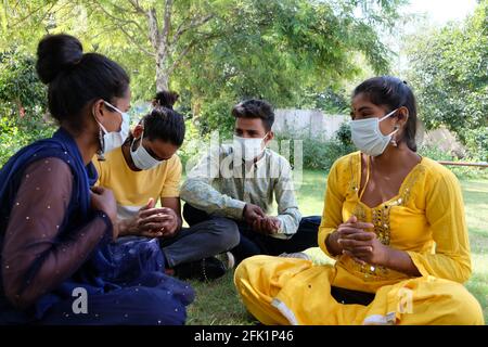 Group of young Indian friends in medical face masks having fun in a park sitting on a grassy ground Stock Photo