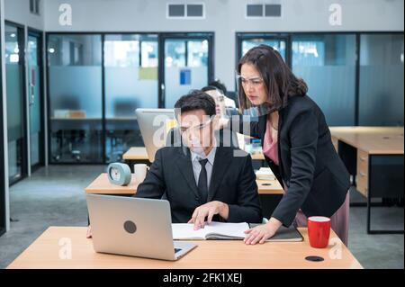Serious asian female executive consulting with male colleague wearing face shield working with laptop at desk in modern office Stock Photo