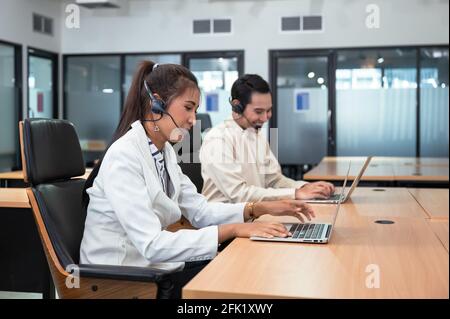 Friendly operator asian woman agent with headset working with laptop on customer consultation in the office with colleague at workplace Stock Photo