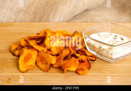 Fresh baked carrot chips on a board with a dipping souse. A blurred sack is background. Homemade chips. Stock Photo