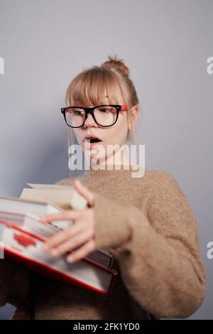 Amazed emotion of nerd caucasian girl. Attractive university or college blonde female student wearing eyeglasses and sweater holding stack of books in hands. Education or knowledge concept Stock Photo
