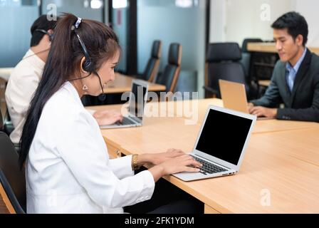 Friendly operator asian woman agent with headset working with laptop on customer consultation in the office with colleague at workplace Stock Photo