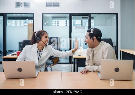 Friendly operator asian colleague agent with headset and laptop talking gossip between working in call centre Stock Photo