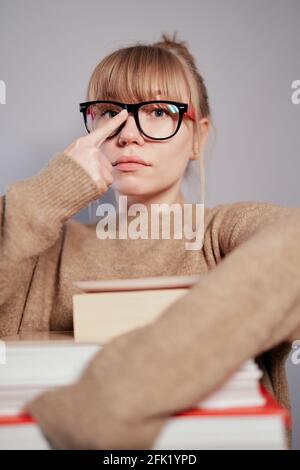 Attractive female caucasian university student wearing beige sweater holding stack of books in hands and correcting her eyeglasses. Nerd, education or knowledge concept. High quality vertical image Stock Photo
