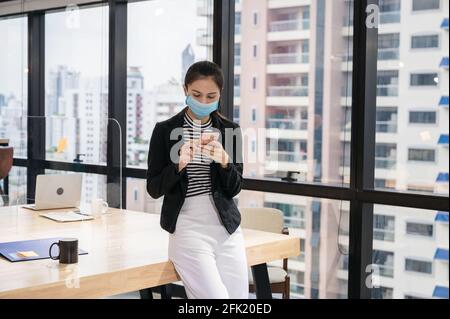 Beautiful young asian woman wearing face mask using smartphone at wooden desk in new normal office. During the outbreak of Coronavirus, Covid-19 Stock Photo