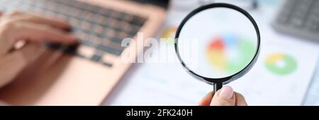 Woman holding magnifying glass in hands over documents with graphs and charts and working on laptop Stock Photo
