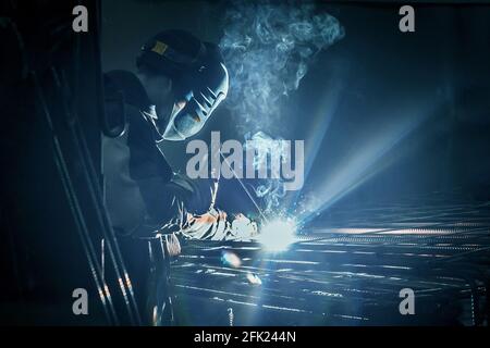 A masked working man is doing welding work on metal structures in a factory or industrial enterprise. Stock Photo