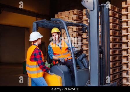 Man worker sitting in industrial forklift at warehouse talking to woman co-worker Stock Photo