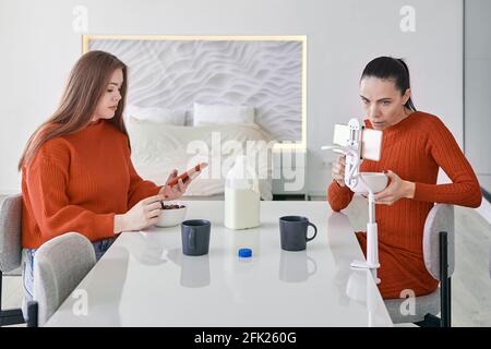 Two women are having breakfast at dinner table in kitchen and browsing information on their smartphones. Stock Photo