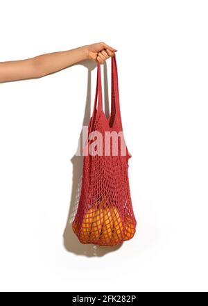 Woman hand holding red string shopping bag with vegetables and fruits in front of white wall background. Eco lifestyle. No more plastic Stock Photo