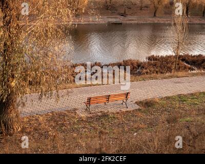 Empty wooden bench on the bank of a narrow water canal. Paths along stream in a magical autumn park with golden foliage in the city. Stock Photo