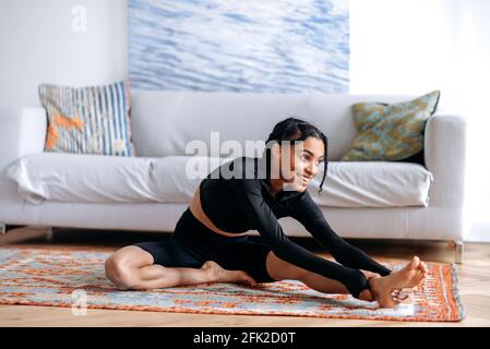 Satisfied young African American woman in black sportswear, doing fitness in living room on carpet, stretching her arms to her feet, doing back stretching, leads healthy lifestyle, looking away, smile Stock Photo