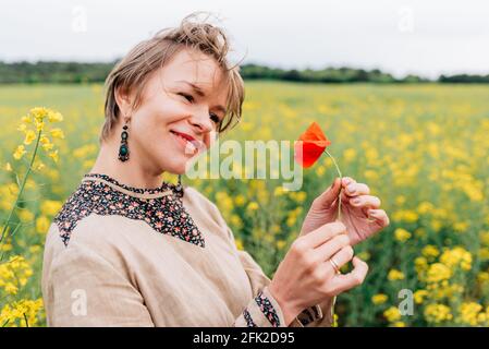 horizontal portrait of an attractive young woman on a flower field. She is smiling while holding a poppy flower Stock Photo
