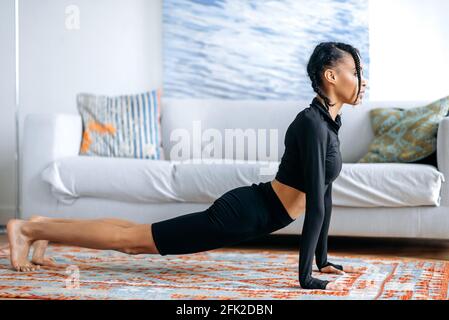 Attractive active slim African American girl in black sportswear, doing stretching at home on the floor, is engaged fitness, leads a healthy lifestyle, care about health