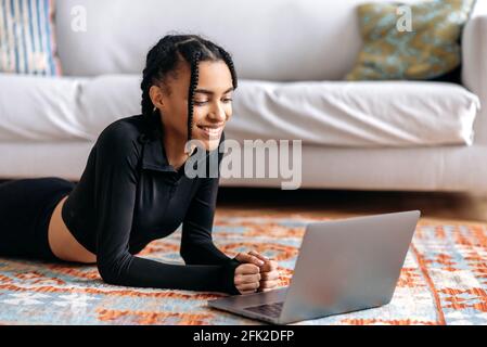 Happy young African American woman with good figure, in sportswear, lies on floor at home, uses laptop, watches video fitness or yoga lessons, goes in for sports, leads healthy lifestyle, smiles Stock Photo