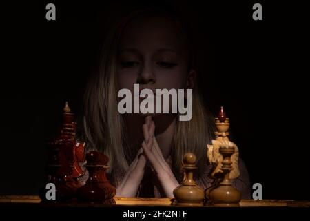 the girl folded her arms and looks thoughtfully at the chessboard against a dark background. old chess. business concept. Stock Photo