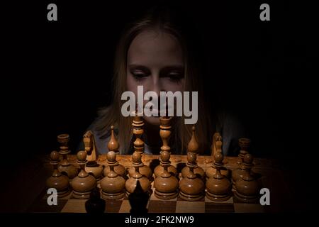 the girl looks thoughtfully at the chessboard against a dark background. old chess. business concept. Stock Photo