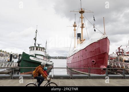 A cyclist with a small child rides past the Arthur Foss tugboat and the Lightship Swiftsure docked at Lake Union Park's Historic Ships Wharf in Seattl Stock Photo