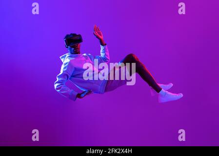 Amazed young African man wearing VR glass headset levitating in the air on futuristic purple cyberpunk neon light background Stock Photo