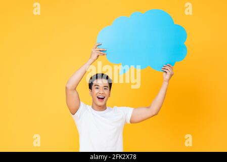 Excited smiling young handsome Asian man looking at camera and holding speech bubble with empty space for text isolated on yellow background Stock Photo
