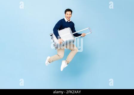 Jumping portrait of young happy smiling handsome Asian tourist man with baggage ready to travel isolated on light blue studio background Stock Photo
