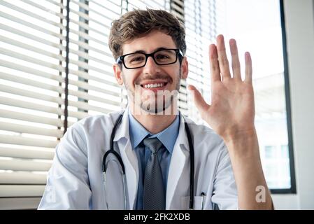 Smiling young male doctor waving hand to camera greeting patient online via distance video calling from the office Stock Photo