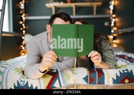 Middle aged couple reading a book in bed, hiding their faces behind the cover. Lying next to each other on a blanket under a garland. Stock Photo