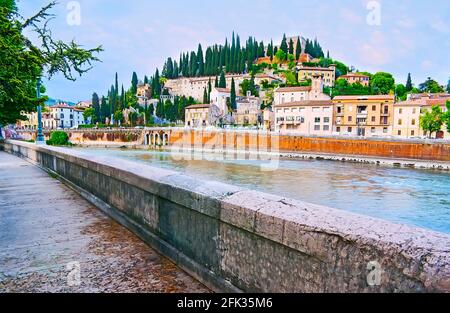 Embankment of Adige River overlooks the San Pietro Hill with tall cypress trees, medieval villas, former San Girolamo Convent and San Pietro Castle at Stock Photo