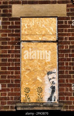 On a boarded up window set in a red brick wall there is stencil graffiti of a man and two flowers which have the anarchy symbol at their centre. Stock Photo