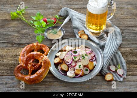 Hearty Bavarian salad with cut veal sausages, roasted pretzel slices on a tin plate, served with a mustard dressing and a mug of lager beer Stock Photo