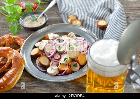 Hearty Bavarian salad with cut veal sausages, roasted pretzel slices on a tin plate, served with a mustard dressing and a mug of lager beer Stock Photo
