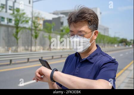 Middle-aged Asian man wearing a face mask and looking at a smartphone in the driveway. Stock Photo