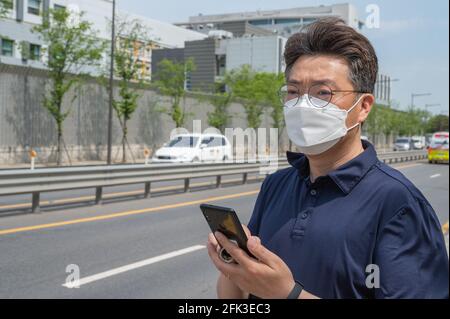 Middle-aged Asian man wearing a face mask and looking at a smartphone in the driveway. Stock Photo
