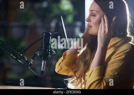 Side view close up of attractive young woman in modern headphone singing into black microphone. Concept of recording trendy songs in music professional studio.  Stock Photo