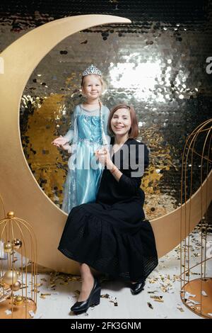 Happy young mother and daughter in beautiful outfits are sitting against the background of a golden wall and looking at the camera Stock Photo