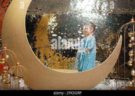 A little girl in the role of a princess rejoices in flying confetti on a golden background. Place for your text. Stock Photo