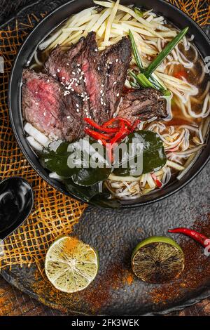 Asian noodles in broth with slow cooked Beef with herbs plated on black background, decorated with spices, lime and chilli peppers Stock Photo
